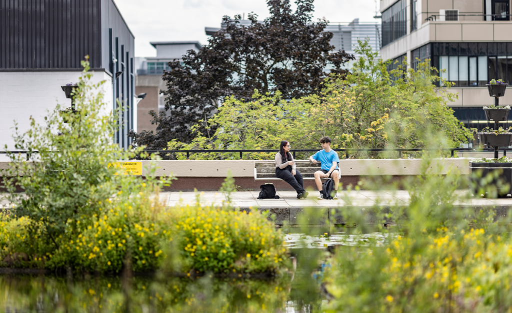 Two people talking on a bench next to Roger Stevens' pond